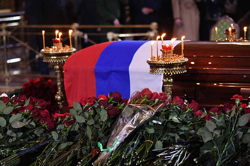 В центре собора стоит закрытый гроб покрытый флагом России. Люди приносят цветы, ставят свечи за упокой. 