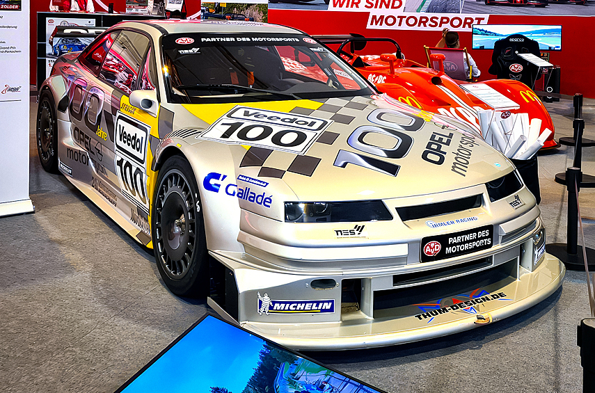 На стенде команды Irmler Racing — гоночный Opel Calibra V6 4x4. Такие машины ездили в немецкой серии DTM, а потом в международной ITC в эпоху технического беспредела «Класса 1», с 1993 по 1996 год. Это Calibra так называемой «Фазы 3», появившаяся в 1994 году. Она ещё сохраняла кое-какие черты серийной машины — например, схему передней подвески «Макферсон» (но только в общих чертах). Под капотом продольно установлен атмосферный V6 2.5 Cosworth KF с сухим картером, который развивает 457 л.с. и «крутится» до 12000 об/мин. Разработанная специально для этой машины трансмиссия Xtrac — изощрённейшая: здесь секвентальная коробка и система полного привода с электро-гидравлическим управлением межосевым дифференциалом, а момент к передним колёсам подводится при помощи бортовых редукторов, которые с обеих сторон «обхватывают» двигатель. Большинство панелей кузова и отделка кокпита выполнены из углепластика — в те годы этот материал широко использовался разве что в Формуле-1. В наше время машина иногда появляется на исторических соревнованиях в Германии.